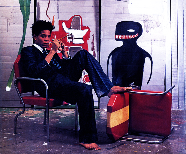 Jean-Michel Basquiat reclines on a chair with one of his creations in the background